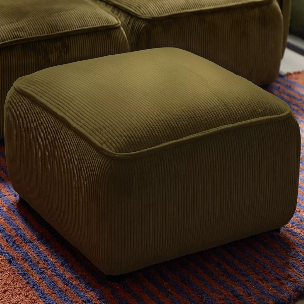 A square, olive green corduroy ottoman from the Macy Modular Sofa collection sits on a textured red and blue striped rug. The ribbed texture of the ottoman's fabric gives it a plush and comfortable look, perfectly complementing the cozy aesthetic of the room.