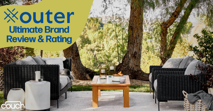 A scenic outdoor seating area featuring two black wicker couches with beige cushions, a wooden coffee table with candles and decor, all surrounded by trees and greenery. Text on the top left reads "Outer Ultimate Brand Review & Rating" with a logo at the bottom left corner.