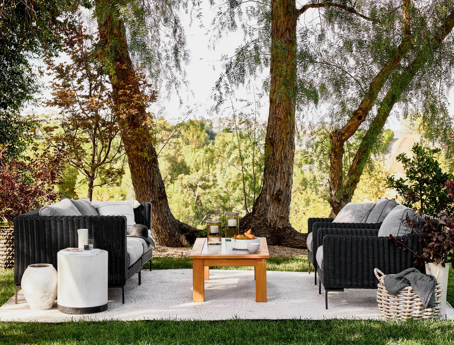 A serene outdoor seating area features two black wicker sofas with light cushions facing each other across a wooden coffee table. The setup is shaded by two large trees and overlooks a lush, green landscape. A white side table and a woven basket are also present.