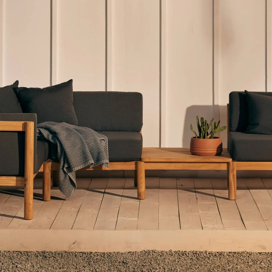 A minimalist outdoor seating area featuring two black cushioned wooden lounge chairs with matching pillows. A small wooden coffee table between the chairs holds a terracotta pot with a green cactus. A grey blanket is draped over the arm of one chair.