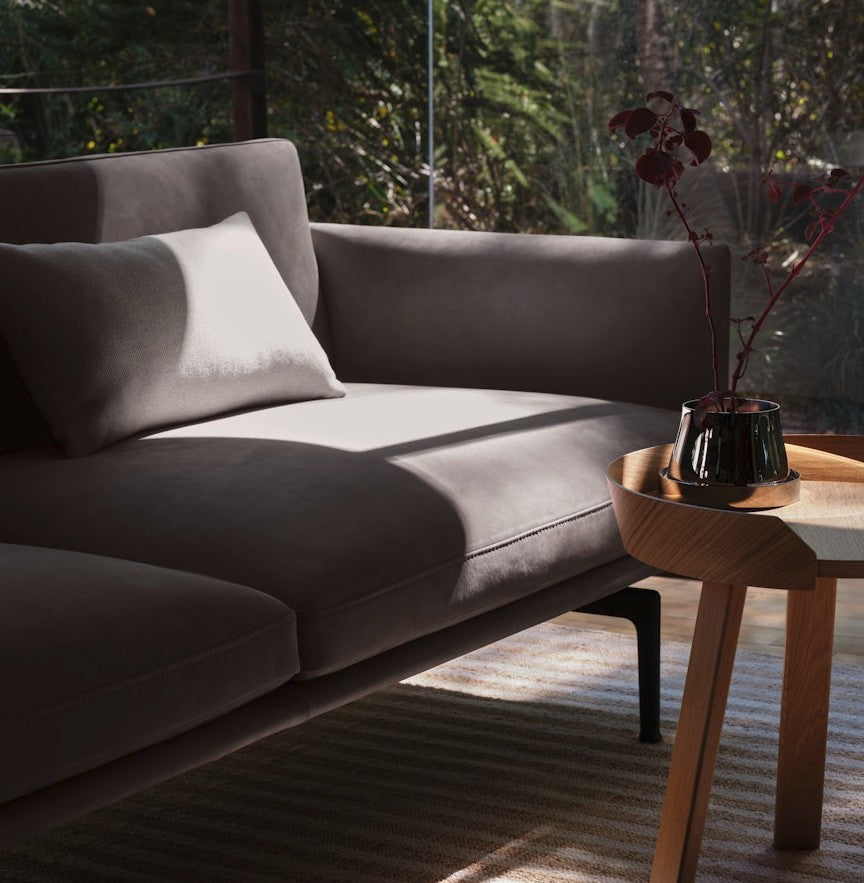 A modern living room with the Outline Sofa 3 Seater in dark gray bathed in sunlight, complemented by a round wooden side table. A dark vase adorned with minimalist floral branches rests on the table. The scene exudes tranquility, with a view of lush greenery visible through the glass window.