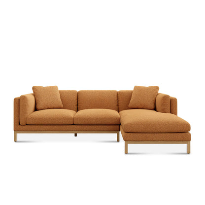 The Owen Chaise Sectional Sofa is a contemporary, L-shaped tan sectional with a chaise lounge on the right side. It boasts a textured fabric finish and comes decorated with two matching cushions placed on the left. The sofa is supported by elegant wooden legs.