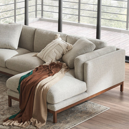 The Owen Chaise Sectional Sofa, a modern beige sectional with cushions and a geometric-patterned pillow, is situated in a sunlit room. A brown and green throw blanket is elegantly draped over one corner. Large windows allow for a view of the wooden deck and surrounding trees outside.