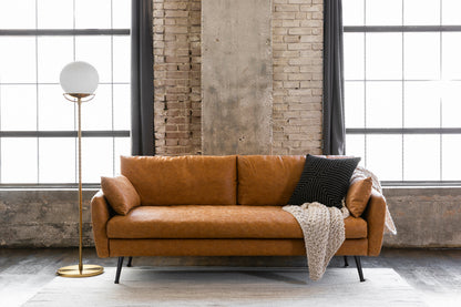 A modern living room features the Park Sofa in distressed vegan leather, decorated with a black pillow and cream knit blanket, situated in front of expansive industrial windows. To the left stands a tall, gold floor lamp with a white globe light.