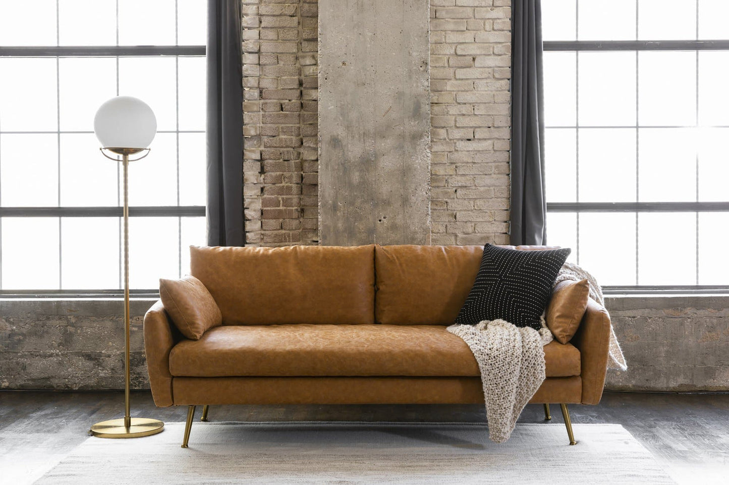 A stylish room features the Park Sofa in distressed vegan leather, decorated with black and beige pillows, set against a brick wall. Large windows flood the space with natural light. A modern floor lamp is positioned to the left, and a textured rug adds warmth to the dark wooden floor.