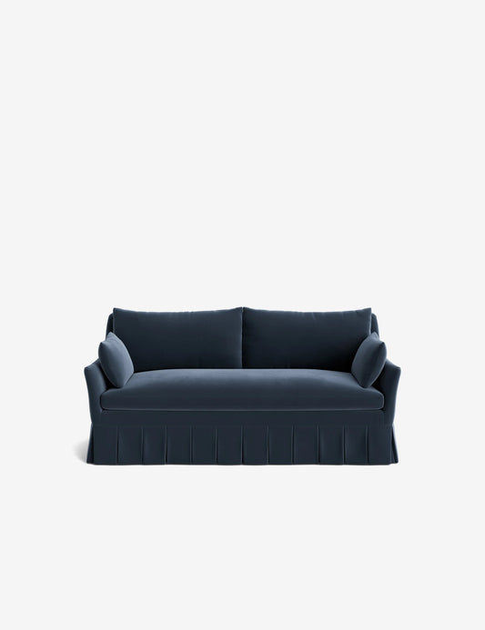 The Portola Pleated Sofa, featuring a dark blue hue and modern design, showcases two plush back cushions, armrest cushions on each side, and a pleated skirt at the base against a simple white backdrop.