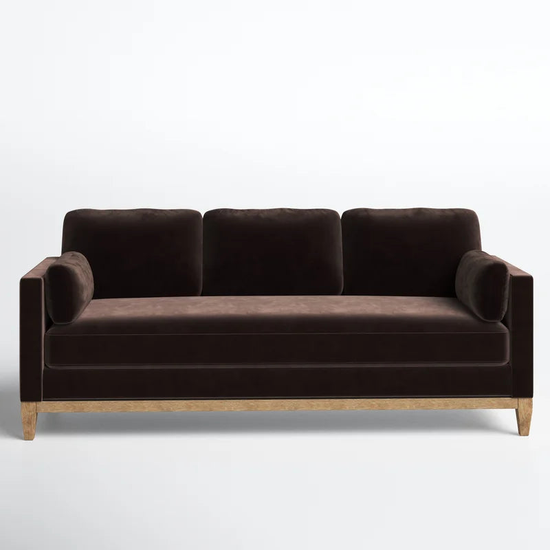 The Pera 84" Upholstered Sofa, featuring a contemporary design with dark brown velvet upholstery, wooden legs, and three cushions, is set against a plain light gray backdrop.