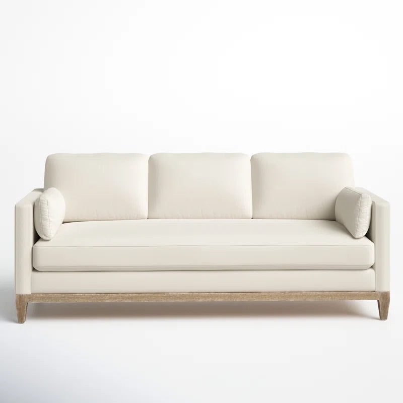 The Pera 84" Upholstered Sofa is a modern, minimalist piece in a cream color, featuring three cushions and two bolster pillows with a simple wooden base, beautifully displayed against a plain white background.