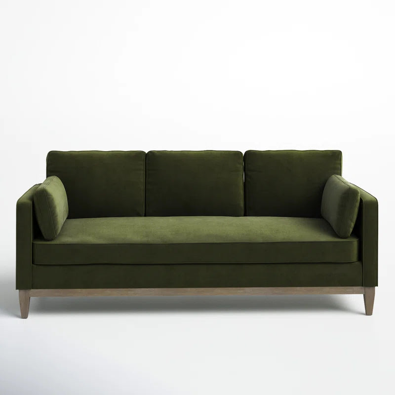 The Pera 84" Upholstered Sofa is a modern piece in dark green velvet, featuring three back cushions and two side pillows. It showcases a sleek design with wooden legs, all set against a plain white background.