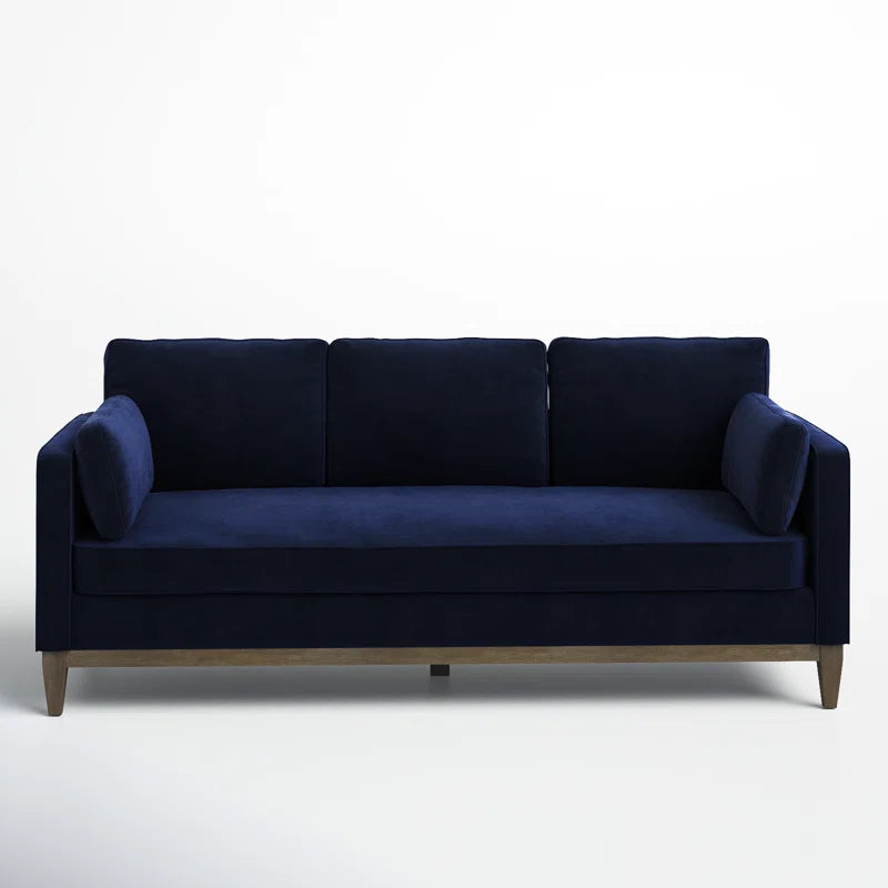 The Pera 84" Upholstered Sofa is a navy blue velvet piece featuring three back cushions and two side pillows, supported by wooden legs and set against a plain white background.