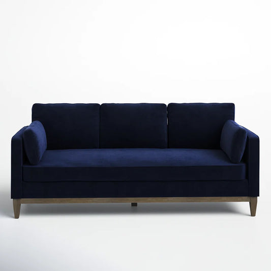 The Pera 84" Upholstered Sofa is a navy blue velvet piece featuring three back cushions and two side pillows, supported by wooden legs and set against a plain white background.