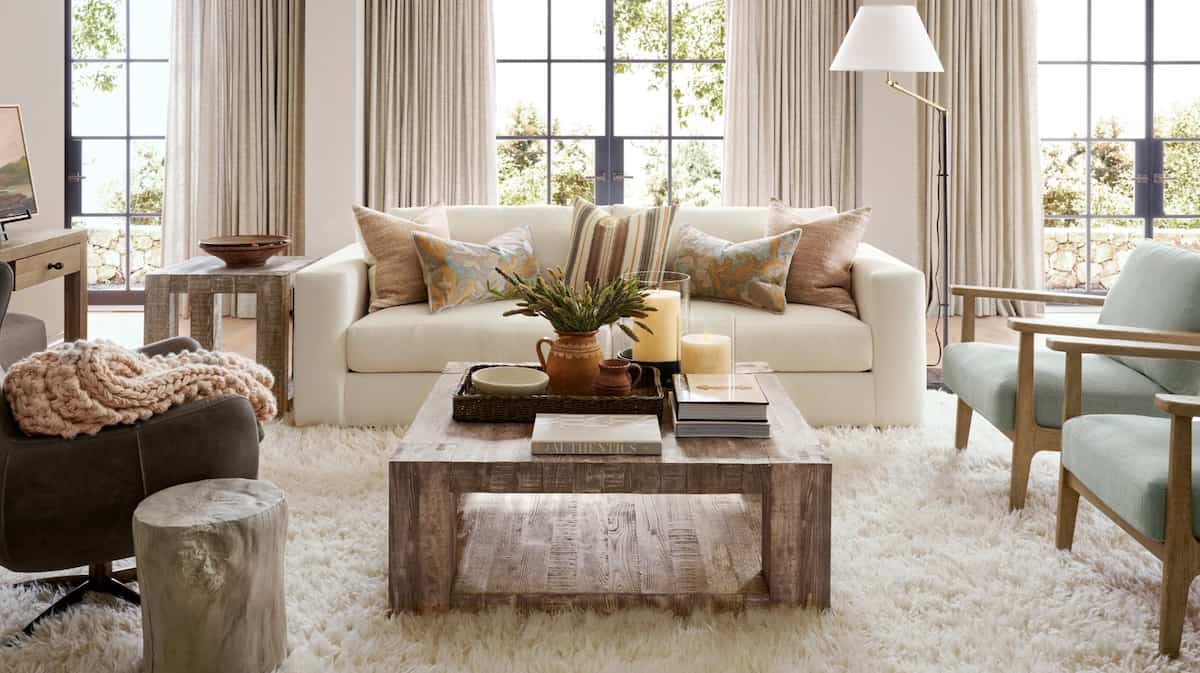 A cozy living room with a beige sofa adorned with pillows, a wooden coffee table holding candles and a plant, and two light green chairs. Large windows with light curtains and a lush, cream-colored rug complete the inviting space.