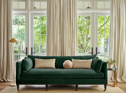 A stylish living room features the Margot Velvet Sofa in dark green, adorned with beige cushions. Large windows behind the sofa allow natural light to flood in, flanked by tall cream curtains. On the left stands a gold floor lamp, while a small side table is positioned on the right.