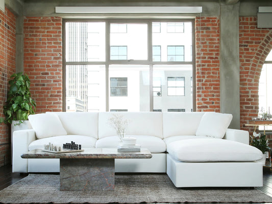 A modern living room showcasing a spacious 3-Seat Modular Chaise Sectional is set against a brick wall with large windows. A rectangular coffee table, adorned with a chess set and decorative plant, rests on a textured rug, while additional plants enhance the corners of the space.