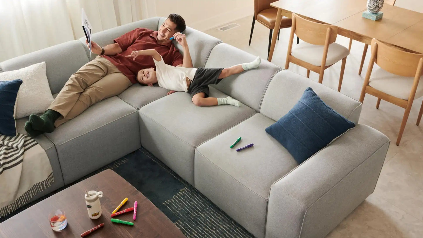 A father and child lounge on the spacious Mambo Loveseat, colored in a light gray. The father reads a book while the child playfully lies nearby, surrounded by colorful markers. A dining table with chairs can be seen in the background.