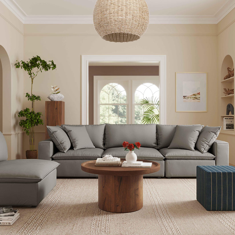 A cozy living room features the Cali Performance Fabric Modular 4 Piece Sectional in gray, paired with a round wooden coffee table. A large wicker pendant light hangs overhead, while decor includes a potted plant, an assortment of books, and a vase filled with red flowers. Neutral tones and natural light contribute to the warm atmosphere.