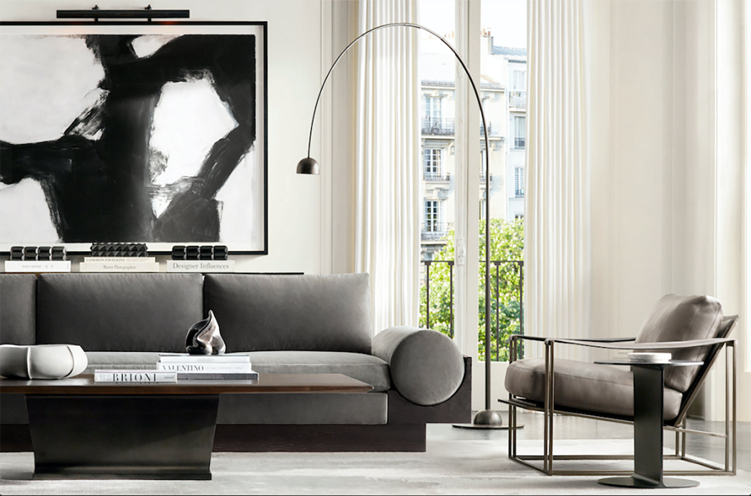 A modern living room featuring a grey sofa, wooden coffee table with books and decor, and a metal-framed armchair. A black and white abstract painting hangs on the wall, and a floor-to-ceiling window with white sheer curtains offers a view of buildings outside.