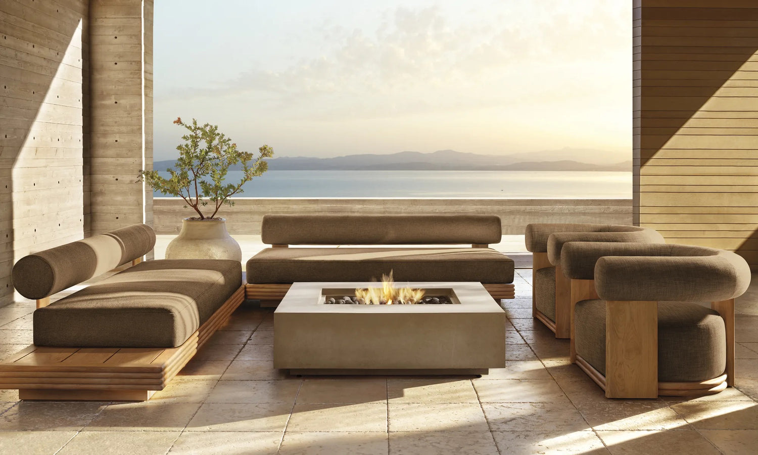 A cozy patio with modern furniture overlooks a serene body of water. The seating arrangement includes cushioned sofas and chairs around a square fire pit. A potted plant adds a touch of greenery, and the setting is bathed in soft, warm light.