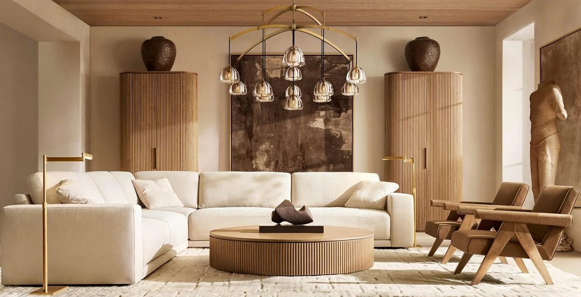 A modern living room features a large, curved white sectional couch with matching pillows, flanked by two wooden cabinets with vases on top. A round wooden coffee table sits at the center, with contemporary chairs and a brass chandelier above. Neutral-toned decor completes the space.