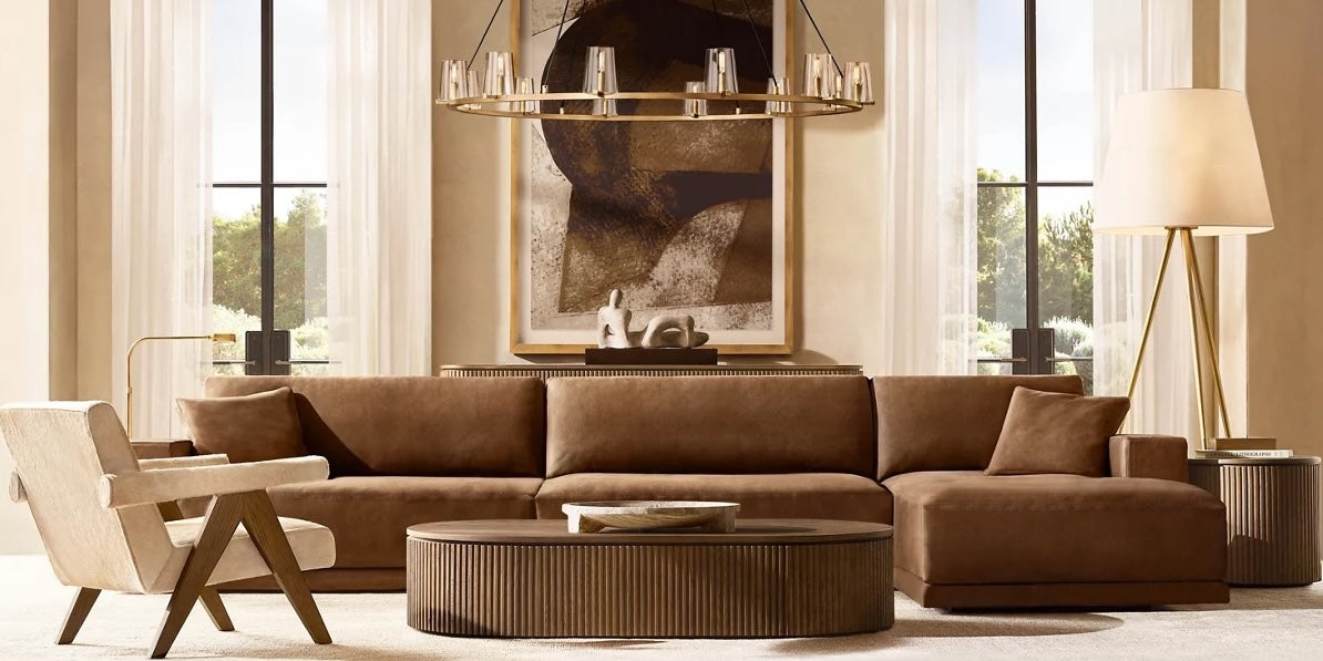 A modern living room with a large brown sectional sofa, a beige armchair, and a round coffee table. A contemporary chandelier hangs from the ceiling, and an abstract painting adorns the wall. Two large windows let in natural light, and a tall floor lamp stands to the right.