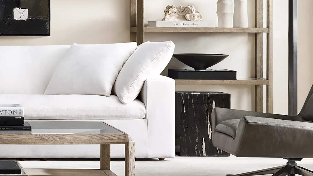 A modern living room featuring a white sofa with a matching pillow, a glass-top wooden coffee table, and a dark gray armchair. Behind the sofa is a wooden shelf with decorative items, including a sculpture, vases, and books.