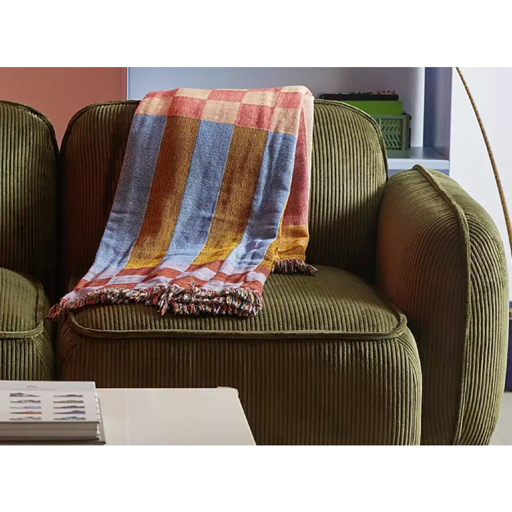 A Macy Modular Sofa in green features a colorful striped throw blanket draped over its back. In the foreground, there's a white table with a magazine, and to the right, a curved lamp is partially visible.