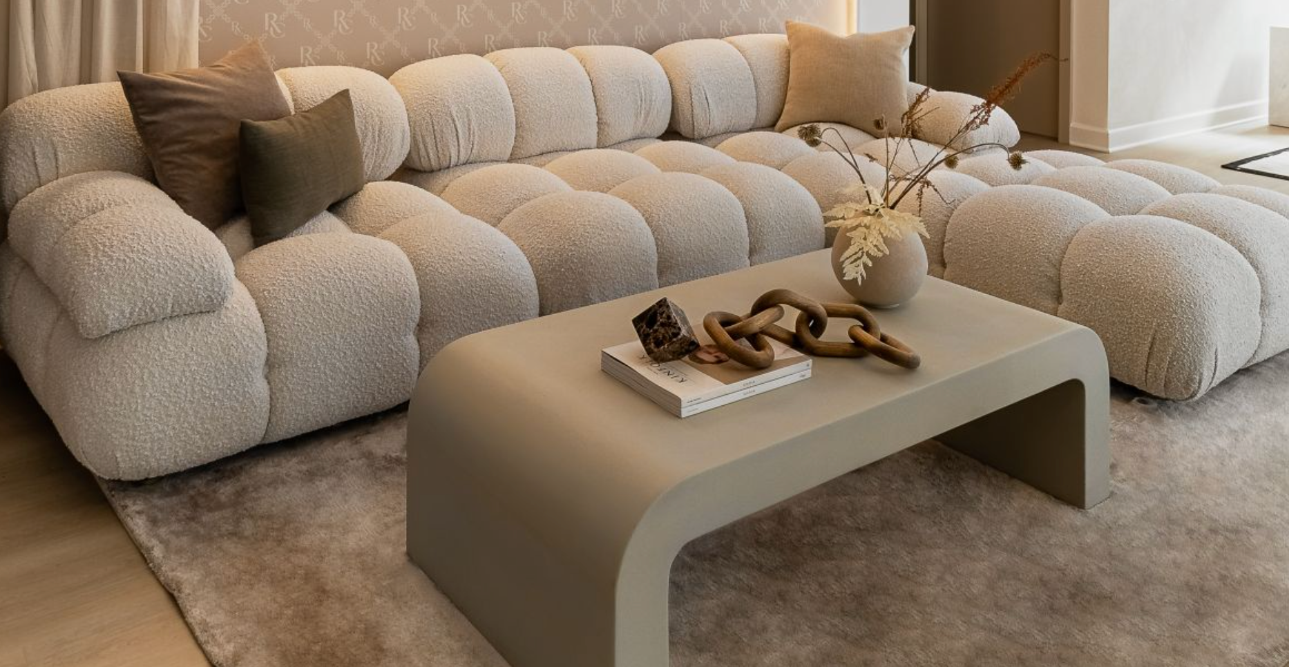 A modern living room features a beige tufted sofa with pillows, a beige curved coffee table, two books, a decorative chain, and a vase with dried flowers. The room has a neutral color palette and a plush beige carpet, creating a cozy atmosphere.