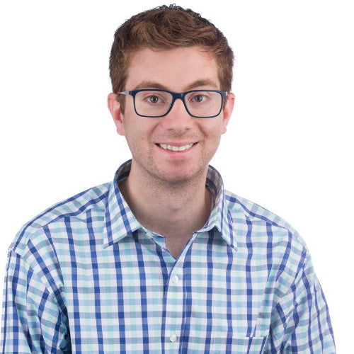 A young man with short brown hair and black-framed glasses is smiling. He is wearing a blue, green, and white checkered button-up shirt. The background is plain white.