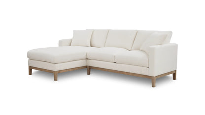 The Any Day Sectional is a white L-shaped sofa featuring wooden legs and plush cushions, complete with a chaise lounge on the left side, all set against a white background.