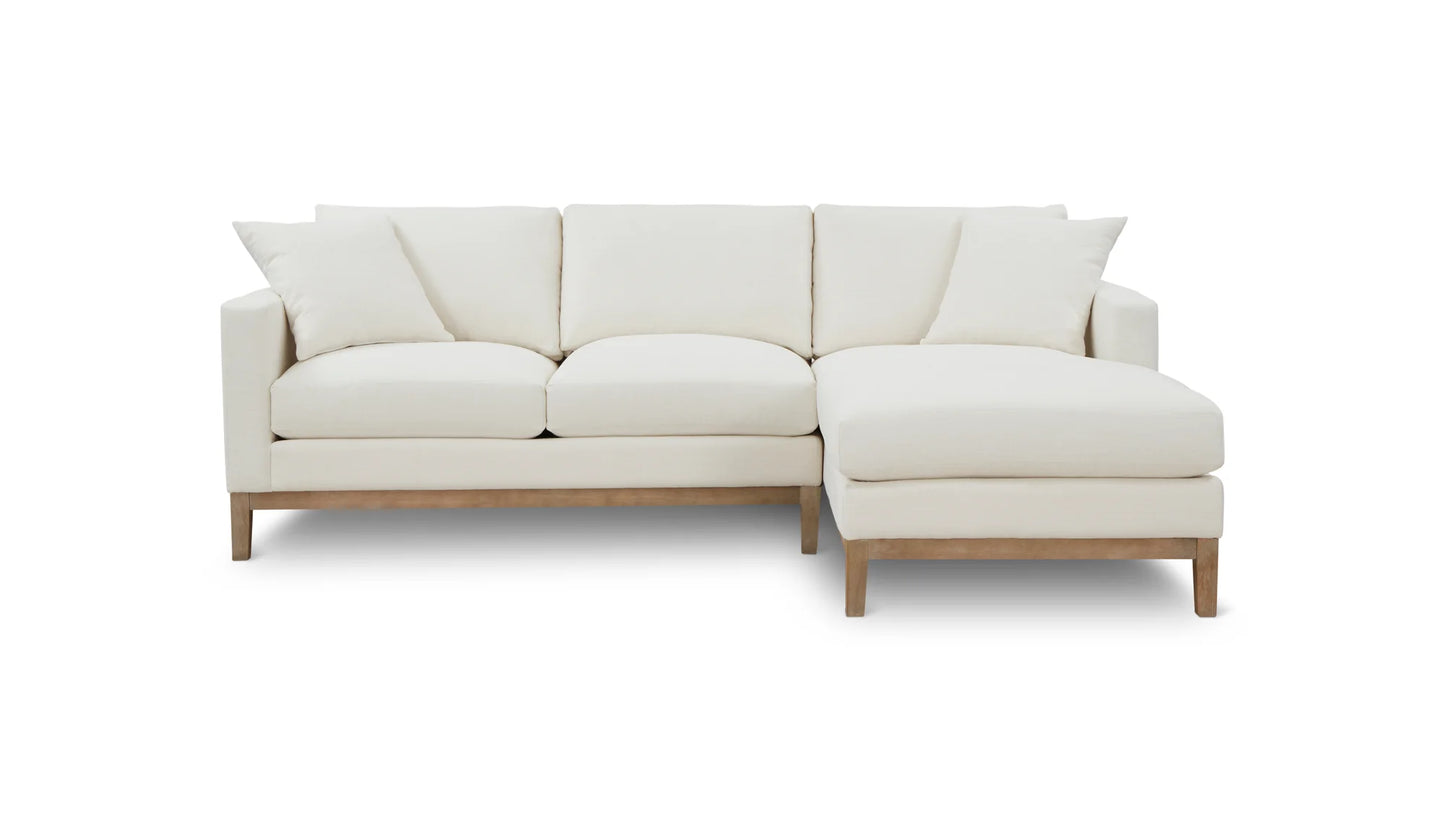 The Any Day Sectional is a contemporary, white sectional sofa with a chaise lounge on the right side. It features plush cushions and wooden legs, designed for a modern living space. The background is plain white.