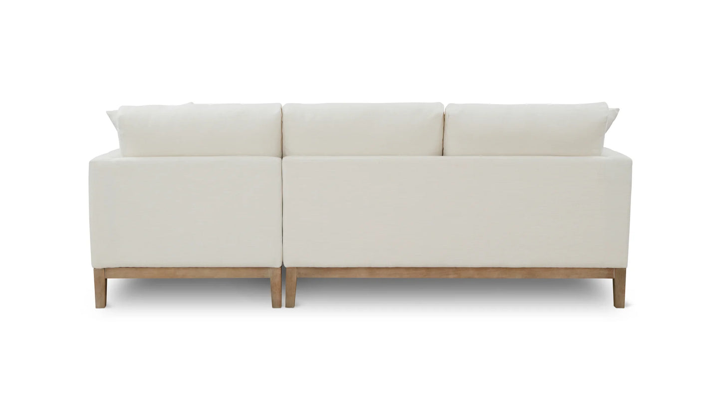 The Any Day Sectional is a minimalist white sofa featuring wooden legs and viewed from the back. It showcases a clean design with three back cushions and a smooth, uninterrupted surface.