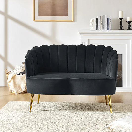 A stylish living room features the Ariel Scalloped Loveseat, which boasts a dark velvet finish, a scalloped back, gold legs, and a cushioned seat. It rests on a light area rug in front of a white fireplace adorned with books, a sculpture, and candles. A framed picture hangs above.