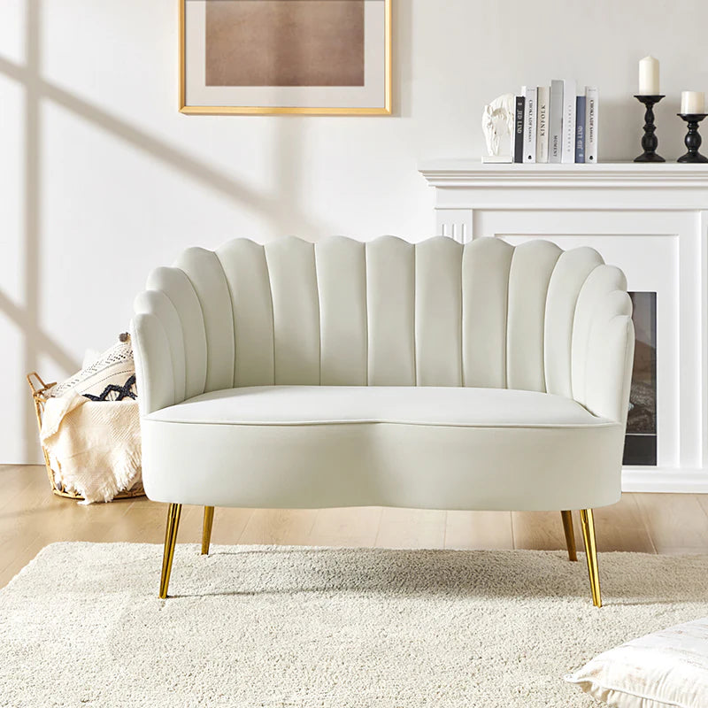 In a bright living room, the Ariel Scalloped Loveseat in light grey, featuring elegant gold legs, is centrally positioned. Behind it lies a cozy setup with a fireplace adorned with candles and stacked books, accompanied by a framed abstract artwork. On the wooden floor rests a soft cream rug.