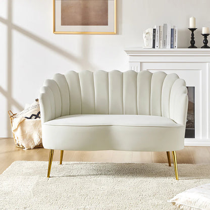 In a bright living room, the Ariel Scalloped Loveseat in light grey, featuring elegant gold legs, is centrally positioned. Behind it lies a cozy setup with a fireplace adorned with candles and stacked books, accompanied by a framed abstract artwork. On the wooden floor rests a soft cream rug.