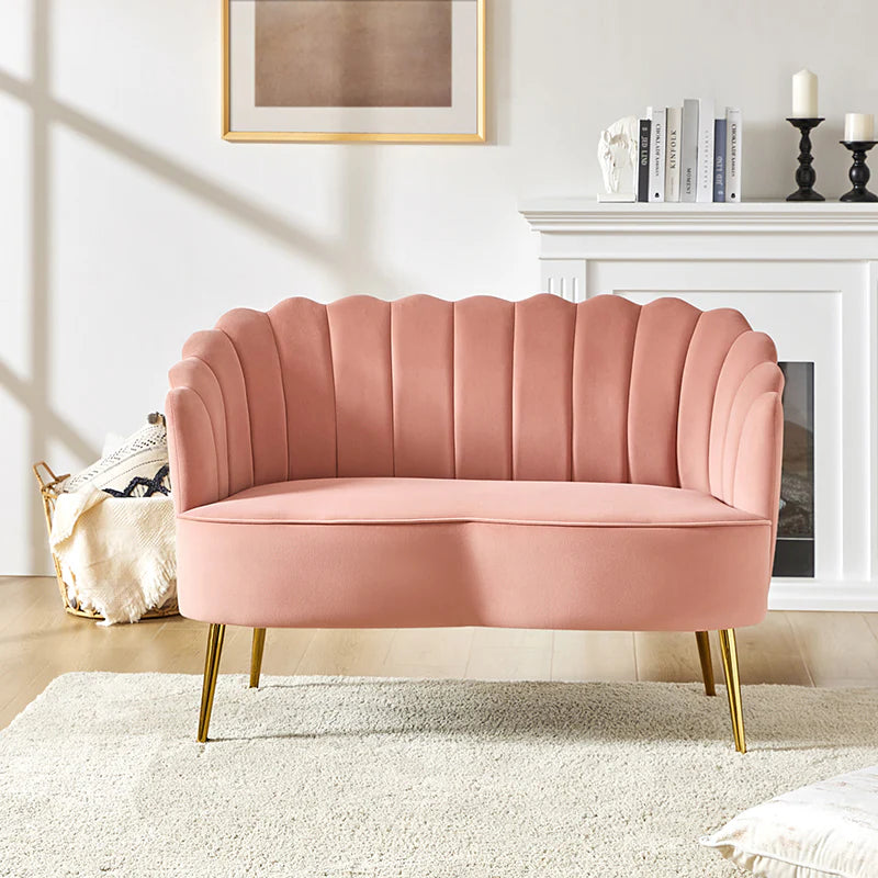 An Ariel Scalloped Loveseat in pink velvet, featuring a scalloped backrest and gold legs, is positioned on a white rug within a living room. In the background, a white mantel exhibits books and candles, while a framed artwork adorns the wall.