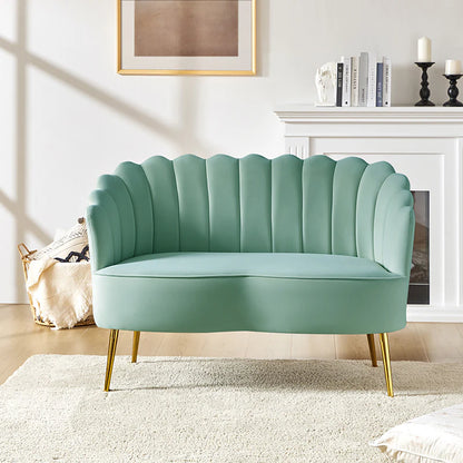 The Ariel Scalloped Loveseat, in a chic mint-green velvet with a scalloped back and gold legs, is placed in a bright living room. It rests on a cream rug before a white fireplace decorated with books, a sculpture, and candles. A large piece of abstract wall art hangs above.