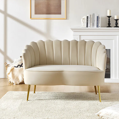 An Ariel Scalloped Loveseat in cream with gold legs sits on a beige rug in a bright living room. In the background, a white mantel above the fireplace holds a framed abstract picture, a stack of books, and candles.