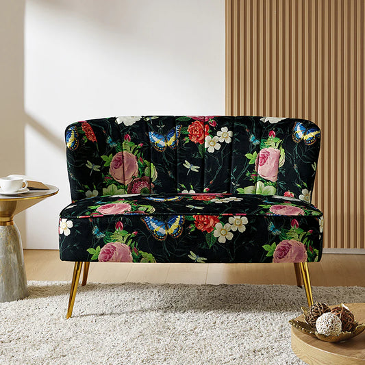 The Coraline Loveseat adds style with its floral pattern on a black background, showcasing vibrant flowers and butterflies. It features elegant gold metal legs and is set in a room with striped wall panels alongside a small side table holding a white teacup. The floor is carpeted, enhancing the cozy ambiance.