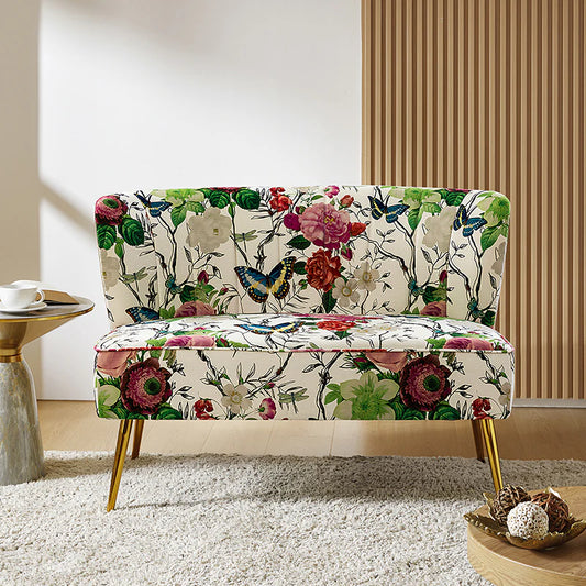 The Coraline Loveseat is a stylish piece adorned with a floral and butterfly print, showcasing vibrant colors such as green, red, and purple. It rests on gold legs and sits atop a light-colored rug next to a small round table. In the background, wooden paneling adds warmth.