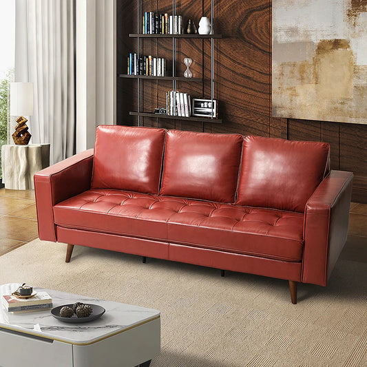 An Anatole Leather Sofa in red, featuring three cushions, is situated in a modern living room. The background includes a bookshelf and a piece of textured wall art. A small coffee table with a decorative centerpiece is placed in the foreground, while light curtains frame the scene.