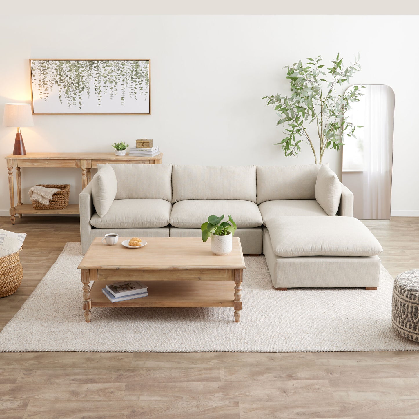 A cozy living room features the Weston Sand Pillow Top 4-Piece L Modular Sectional Sofa, accompanied by a wooden coffee table and a light beige rug. A potted plant and wall art depicting greenery add a natural touch to the space. A lamp and wicker basket sit on the wooden side table, enhancing the warm atmosphere.