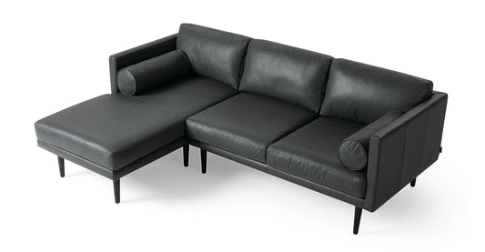 A contemporary Napoli Grey leather sectional sofa, part of the Spectre collection, featuring a chaise lounge on the left side with three cushions and two cylindrical bolster pillows. The sofa showcases sleek, straight arms and black tapered legs, set against a plain white background.