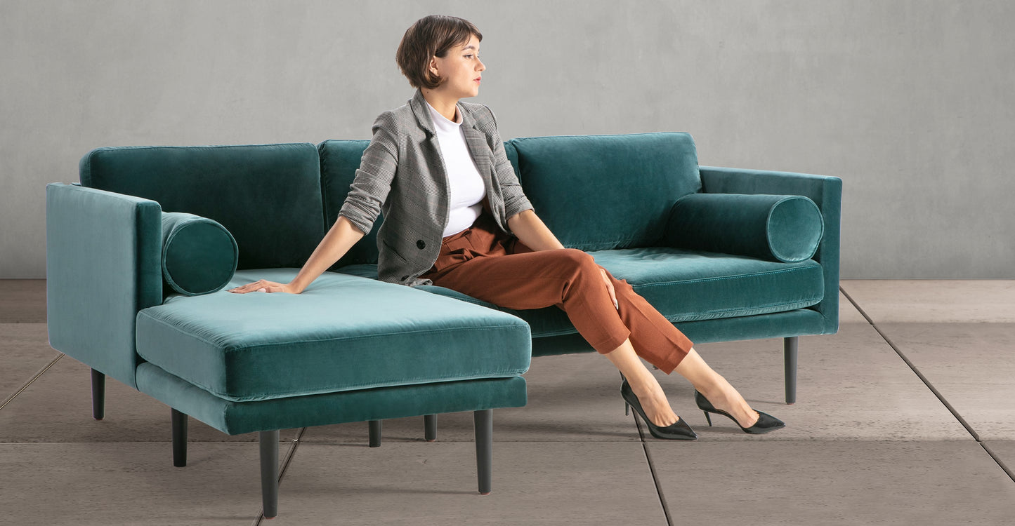 A woman sits on a Napoli Grey Spectre 81" Leather Sofa Sectional Left in a modern, minimalist room. She wears a gray blazer, white top, and rust-colored pants with black heels. The concrete floor and gray wall provide a neutral backdrop for the stylish furniture.