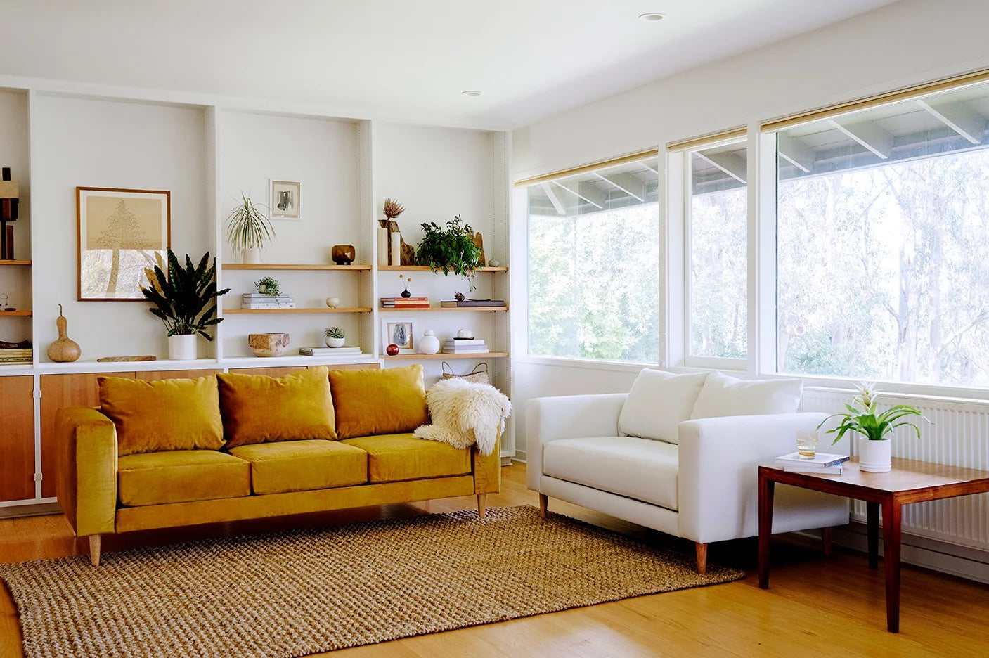 A bright, modern living room with a mustard-yellow couch and a white armchair on a jute rug. A wooden side table with a potted plant sits next to the armchair. Shelves with books, plants, and decor adorn the white walls. Large windows let in ample natural light.