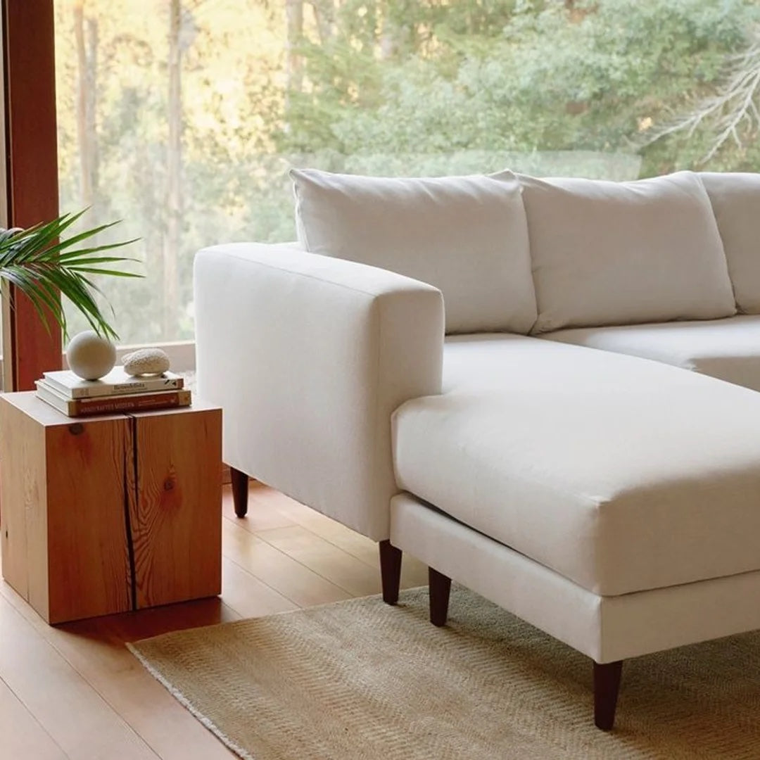 An inviting living room features a white sectional sofa with clean lines, positioned near a large window showcasing a forest view. A wooden side table holds decorative items, including a plant, atop a light-colored rug that complements the serene ambiance.