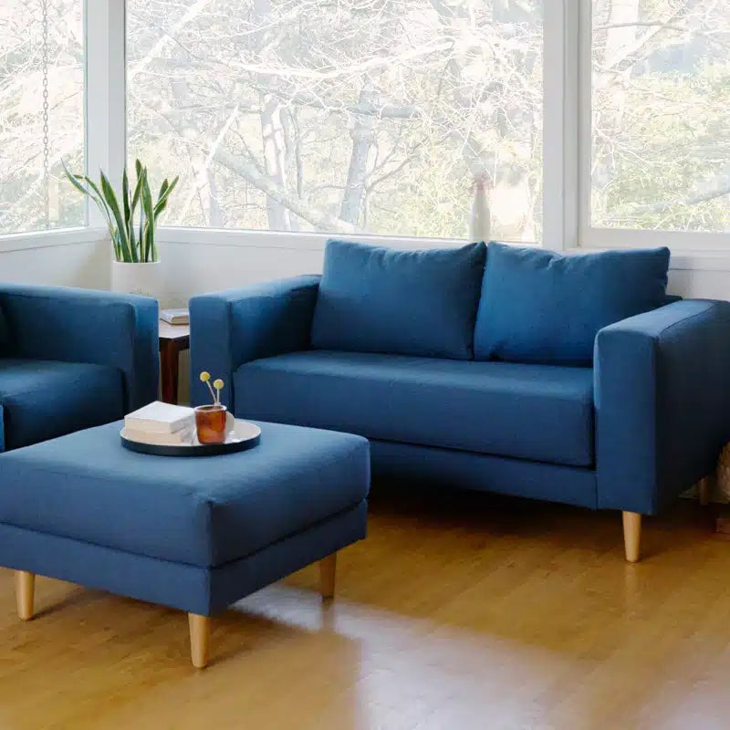 A cozy living room featuring a blue loveseat, matching armchair, and a square ottoman with a tray holding a cup and book. The furniture has wooden legs and is set against large windows, letting in natural light and offering a view of bare trees outside.