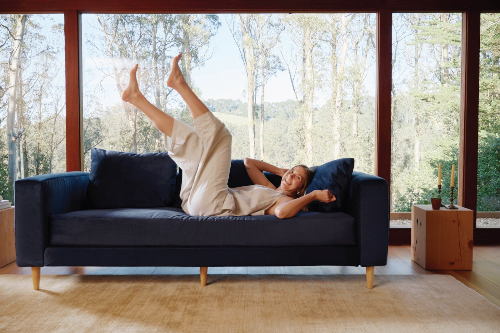 A woman wearing a cream-colored outfit is lying playfully on a navy-blue sofa with her legs raised. She is smiling, and large windows behind her showcase a scenic view of trees and a hillside. Candles and a small plant are on a wooden box to the right of the sofa.