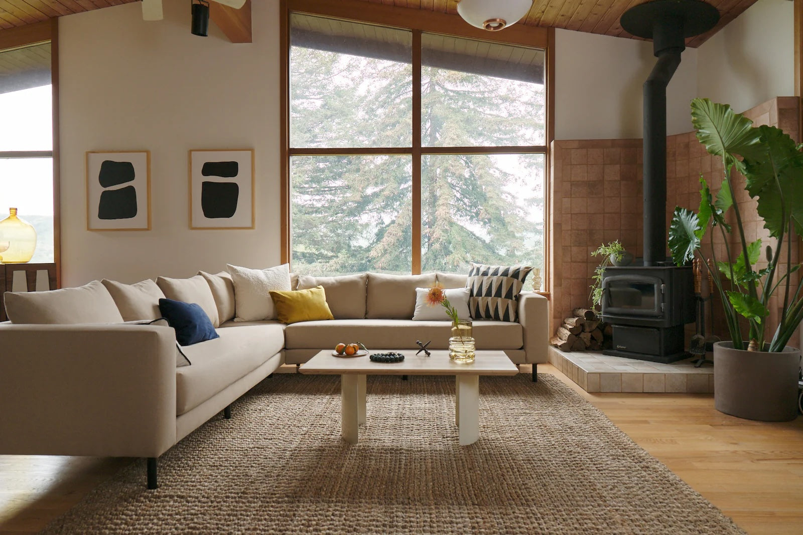 Spacious living room with a modern, neutral-toned sofa, assorted throw pillows, a wooden coffee table, and a wood-burning stove in the corner. Large windows showcase an outdoor view of trees. Minimalist art and indoor plants add to the cozy, contemporary ambiance.