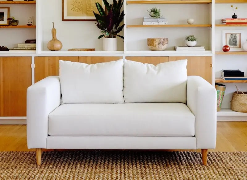 A modern living room featuring a white two-seater sofa with wooden legs. Behind the sofa, there is a white and wood-paneled bookcase with various decorative items, including plants, books, and vases. A woven rug lies on the wooden floor in front of the sofa.