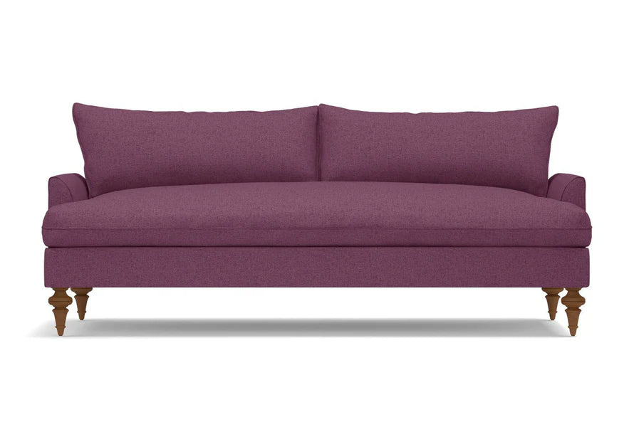 The Saxon Sofa is a modern piece in purple, featuring wooden legs and a sleek, minimalist design with a cushioned backrest. Its smooth fabric texture makes it an ideal choice for a contemporary living room setting.